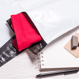 A white poly mailer on a desk, a folded red garment beside a notebook and pen.