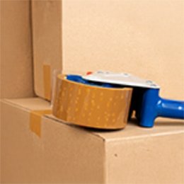 A hand-held tape gun sealing the top seam of a kraft cardboard carton with brown carton-sealing tape.